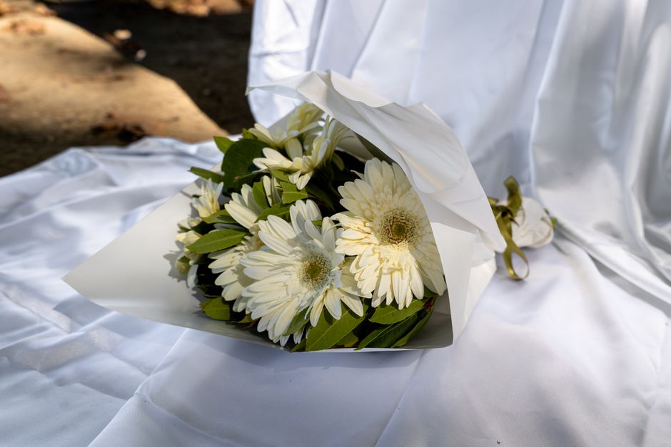 A bouquet of fresh white Gerbera daisies and white chrysanthemums, mixed with green foliage, wrapped in elegant white paper and tied with a golden ribbon, resting on a satin-like white cloth with natu