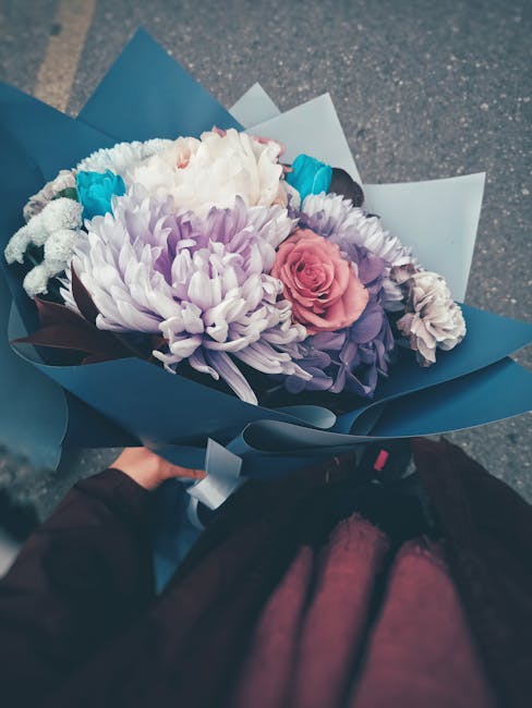 A close-up view of a floral bouquet featuring a variety of fresh flowers, including a large white chrysanthemum, soft pink and purple roses, and small turquoise accent blooms, arranged in a natural, l