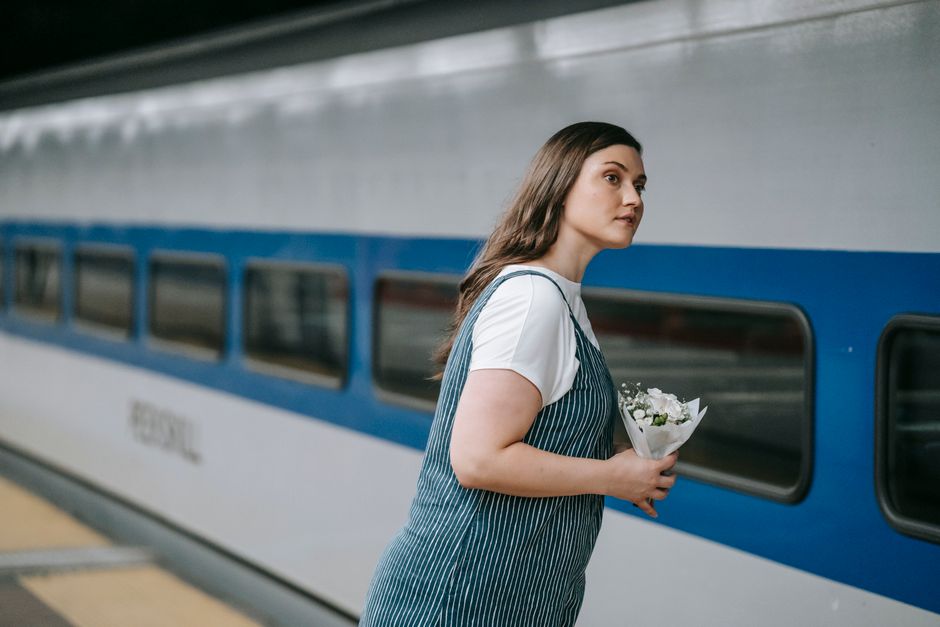 A young woman with long, brown hair, wearing a white t-shirt and a blue-and-white striped apron, stands on a train platform holding a small bouquet of fresh white flowers wrapped in white paper. She a