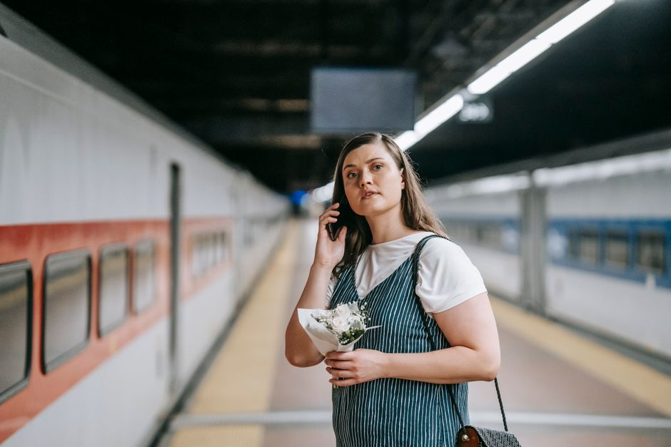 A young woman with fair skin and long brown hair is standing on an underground train station platform, holding a bouquet of white flowers wrapped in clear paper. She is wearing a white t-shirt layered