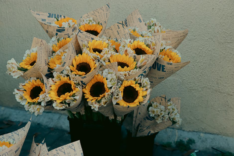 A bouquet of fresh sunflowers with bright yellow petals and dark centers, arranged with white daisies and smaller filler flowers, wrapped in brown paper printed with love-themed words and blue accents