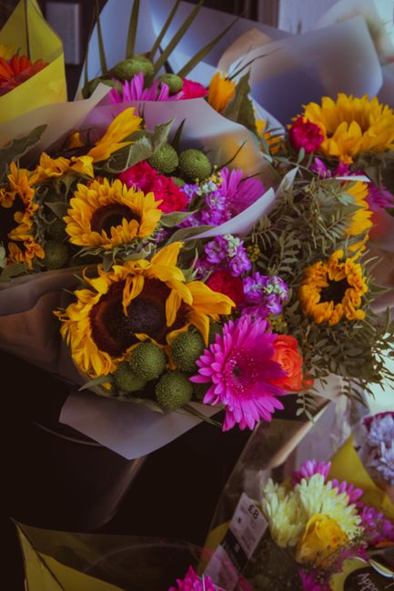 A vibrant bouquet of fresh flowers featuring yellow sunflowers with dark centers, bright pink gerbera daisies, and small green billy balls, complemented by purple and pink blossoms, all arranged with