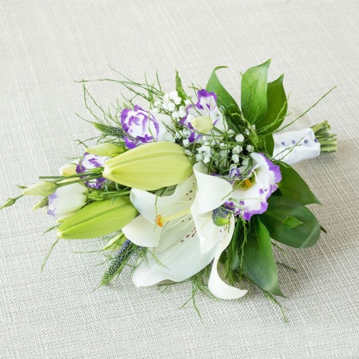 A sophisticated, hand-tied bouquet of white lilies and purple lisianthus stretches diagonally across a neutral, light beige fabric that has a subtle, woven texture reminiscent of natural linen often used on wedding tables in Mitcham venues. The arrangement is distinctly elongated, with the lush head of flowers on the left and the neatly bound stems tapering to the right. Dominating the centre are fully opened white lilies, their broad, velvety petals gently arched back to reveal warm yellow-orange stamens dusted with pollen and faint, russet speckles near the base. Tucked amongst them, several smooth, light green lily buds stand upright, still closed but full of promise, like a quiet pause before the next wave of blooms. Woven through this framework, lisianthus flowers unfurl in soft, ruffled layers: creamy white petals finished with bold, purple edges and some deeper violet centres, creating a striking yet harmonious contrast. Scattered sprigs of delicate white gypsophila add a fine, misty layer, filling gaps with a soft, cloud-like texture that makes the bouquet feel airy rather than heavy. The foliage is beautifully varied, with glossy, dark green leaves offering structure and depth, complemented by slender, wispy greens and a few spikier accents that introduce rhythmic lines and movement, as though the stems had just been gathered from a garden near Ravensbury Park. At the narrow end, the stems are carefully wrapped in a crisp white ribbon adorned with tiny, evenly spaced bead-like details, emphasising that this piece is crafted for a special occasion-perhaps a bridal bouquet, bridesmaid posy, or a significant gift. Gentle, even lighting washes over the flowers, enhancing their fresh textures and true colours without glare, and the simple background ensures all attention rests on the refined beauty and craftsmanship of the floral design.