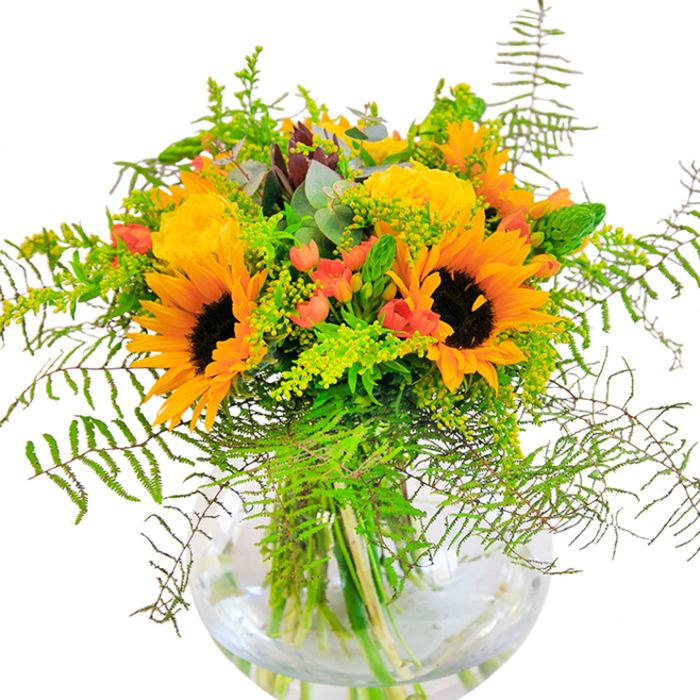 With a first impression like a sunbeam breaking over Mitcham Common, this lush and expressive bouquet radiates warmth and vibrancy. Sunflowers with brilliant yellow petals and velvety dark centers preside over the composition, accompanied by garden roses in soft, buttery gold-each bloom opening in classic, spiraled elegance. Clusters of scarlet Hypericum berries catch the eye, their glossy finish providing a spirited contrast amid the greens. Cascading ferns and luxuriant foliage weave throughout, suggesting the wild beauty of Mitcham's open spaces and the winding paths that invite locals for an afternoon stroll. Slender green spikes, delicate solidago stems with star-like yellow florets, and bluish eucalyptus leaves complete the textural symphony, infusing the arrangement with layered depth and subtle herbal aroma. The round glass vase, reminiscent of the glassblowing craft of nearby South-West London artisans, cradles the stems and reveals their lively green hues, reinforced by clear water within. The bouquet is alive with movement, color, and freshness-a fitting centerpiece for anniversaries, joyful milestones, or simply to bring a sense of celebration into a Mitcham home. Rooted in local character and the artistry of skilled florists, it is a bouquet that invites admiration at every glance.