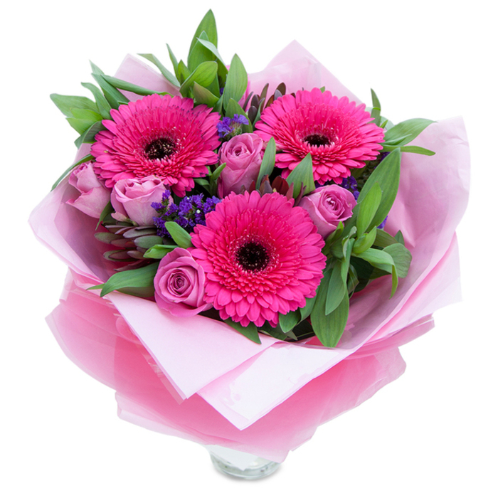 The first thing you notice is the confident pulse of fuchsia from three Gerbera daisies, each with precise, radiating petals around deep, nearly black centres that give the bouquet a graphic, joyful rhythm. Nestled beside them, lavender-pink roses offer a softer cadence: their velvety petals curl inward in a refined, budding shape that invites touch. Scattered throughout are fine sprays of purple statice, adding delicate, papery texture and little pockets of cool colour that create movement across the arrangement. Lush foliage - broad salal leaves and slim eucalyptus stems - forms a verdant scaffold that lifts the flowers and introduces a fresh, slightly resinous scent. The bouquet is lovingly wrapped for gifting with an inner veil of pale pink tissue and an outer sheet of translucent fuchsia-pink cellophane that catches the light and ties every shade of pink together; the folds are deliberate, the edges tucked to present the bouquet like a kept secret. At the base a clear glass vase rim is visible, hinting the flowers are meant to be enjoyed immediately, to sit on a side table or hall console. In my hands as a florist I'm thinking of delivering this to a friend in Mitcham - perhaps to a home on London Road overlooking the common - where its lively colourway would instantly lift a room. The scent is gentle: mostly rose-sweet with a green undertone from the gerberas and eucalyptus. This arrangement reads as an affectionate, celebratory gesture - ideal for birthdays, Mother's Day, or an everyday 'I'm thinking of you' across the neighbourhood.
