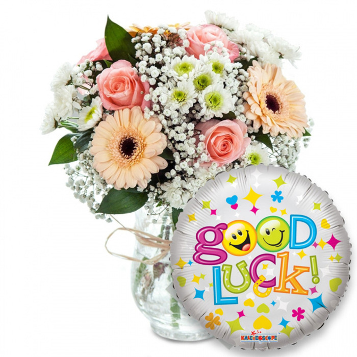This image presents a thoughtfully curated 'Good Luck!' gift, arranged to feel both joyful and reassuring - the sort of bouquet you might pick up on your way past Figges Marsh or from a stall near the town centre. A clear glass vase, modestly tied with raffia or twine, holds a lush composition of blooms: soft, powdery-pink roses whose petals suggest a gentle, lingering scent; warm peach-coloured gerbera daisies with bold, dark brown centres adding sunny contrast; and generous sprays of white baby's breath that float like delicate lace around the main flowers. Interspersed are small white and lime-green chrysanthemum-type spray blooms that introduce a crisp, fresh texture, while deep green foliage frames each blossom and gives the arrangement pleasing depth. In the foreground and slightly right, a shiny silver foil balloon overlaps the vase, its metallic surface catching light and amplifying the festive message Good Luck! in a playful rainbow of letters. The designer detail - the two 'o's in Good transformed into cheeky yellow smiley faces (one winking, one tongue-out) and the tiny decorative stars and hearts scattered across the balloon - injects a pinch of humour and celebration. Rendered on a clean white background with soft lighting, the scene emphasizes the colours and textures: velvety roses, the crispness of chrysanthemum petals, and the airy softness of gypsophila. It's an ideal composition for milestone moments - exams, interviews, farewells or first days - and feels like a small, thoughtful uplift from a local Mitcham hands-on florist.