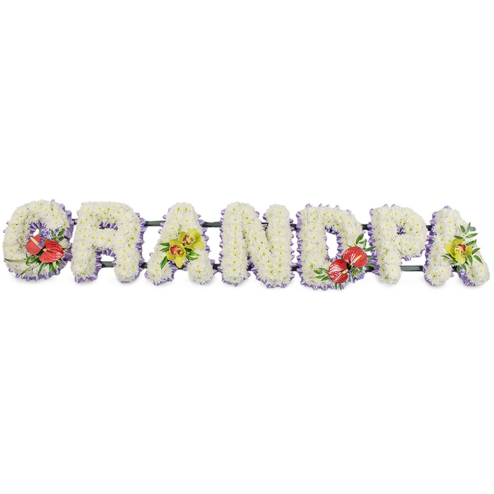 The image presents a long, horizontal floral arrangement spelling out GRANDPA in large block capitals, designed as a classic funeral tribute. Every letter is crafted from a mass of small, pristine white chrysanthemums, the blooms packed so tightly that they form smooth, padded shapes with a gentle, almost velvety texture. The petals catch the light softly, creating subtle shadows that emphasise the contours of each letter and make the word clearly readable from a distance. At the base of the letters, a fine trim of pale lavender ribbon peeks out from under the flowers, outlining each shape in a cool, understated hue that conveys tranquillity and tenderness. The letters are joined and held in place by dark green support bars, which sit mostly behind the flowers; only glimpses of the frame show, hinting at the solid structure beneath the delicate exterior. Colourful accent sprays add personality and heartfelt emotion. On the 'G' and the 'D', striking deep red anthuriums with shiny, heart-shaped spathes and bright yellow spadices nestle amongst rich green foliage, acting as passionate focal points. On the first 'A' and the final 'A', light yellow-green orchids emerge gracefully from the chrysanthemums, their refined petals and speckled throats suggesting grace, admiration, and lasting love. Set against the uncluttered white background, the tribute feels serene and dignified, ideal for placing near the coffin at a Mitcham crematorium chapel or at a graveside service close to Mitcham Common. Created with the eye of an experienced local florist, it offers families a personal way to honour their grandfather's name, expressing sorrow and gratitude through beautifully composed flowers.