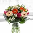 A captivating bouquet designed with Mitcham's unique charm bursts forth in a vivid tapestry of colour and grace. Centred around lush gerbera daisies-two ablaze in energetic orange, two blushing in soft peach-each flower is ringed with a deep brown eye, reminiscent of the diversity and warmth that fills Cricket Green in spring. Tucked snugly alongside are sumptuous magenta roses, their petals tightly furled in an elegant spiral, infusing a sense of depth and affection. Freshness unfurls with snowy white alstroemeria and small clusters of white spray roses, while faint pink blooms and petite white fillers peek from the greenery, adding nuanced layers and volume. The bouquet is beautifully anchored by wide, glossy, paddle-shaped leaves, skillfully curled into a protective, modern collar. Between these, smaller leaves and airy green accents suggest a natural abundance. All is thoughtfully arranged in a clear glass vase, stems standing straight and proud, as if freshly picked on a cool Mitcham morning. Gentle, diffused light from a minimalist backdrop highlights the exuberant colours, allowing the intricate details to take centre stage. Whether gracing a celebratory picnic in Ravensbury Park or conveying heartfelt gratitude to a neighbour, this bouquet embodies the joyful, generous spirit that defines Mitcham and its close-knit community.
